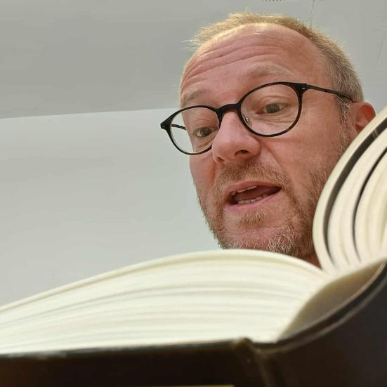 Mand med briller læser højt af bog. Man ser kun toppen og lidt af ryggen af bogen.