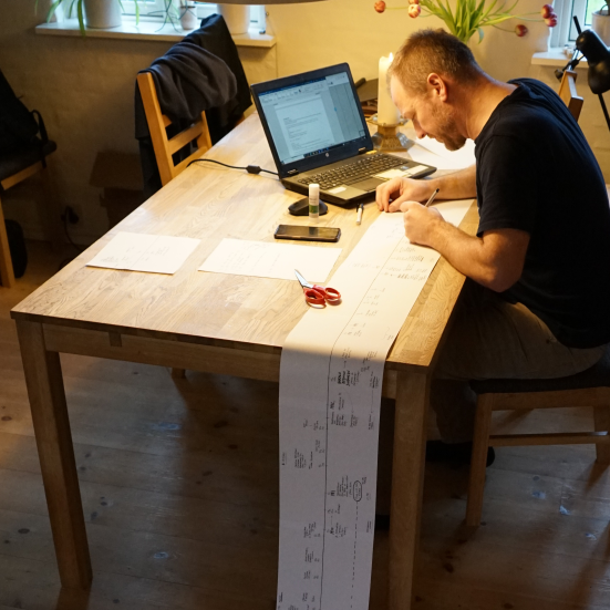 Hans Wendelboe Bøcher sidder ved en bærbarcomputer på et spisebord. Fra bordet løber en flere meter lang papirliste med håndskrevne notater ned og videre ad gulvet.