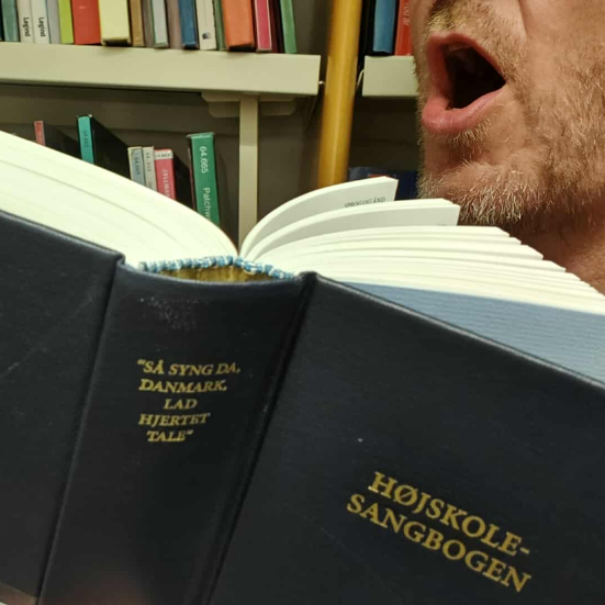 Nærbillede af Højskolesangbogen, med modificeret tekst på ryggen: "Så Syng Da, Brønderslev, Lad Hjertet Tale". I baggrunden mand, der synger og reol med bøger.