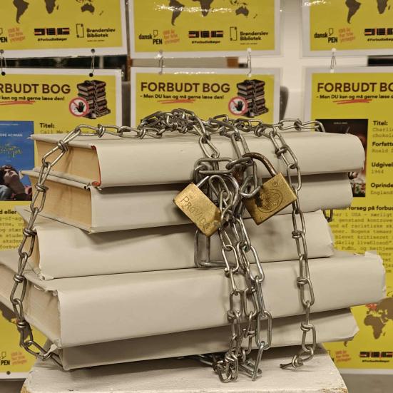 Stak af titelløse bøger, lænket sammen med jernkæder og hængelåse. I baggrunden hænger gule plakater med titler og tekster om bøger, der er, har været eller været forsøgt gjort forbudt. er  