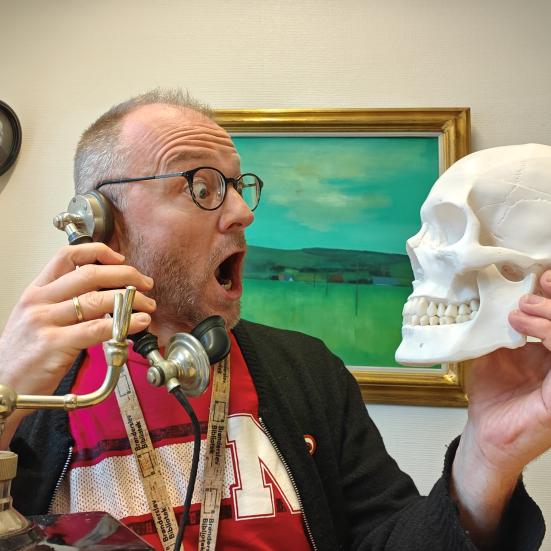 Kultur- og historieformidler Hans Wendelboe Bøcher i rød t-shirt sidder med et menneskekranium i den ene hånd og et gammelt telefonrør i den anden. Han er overrasket ud. I baggrunden hænger et grønt maleri og et portrtæt af N.F.S. Grundtvig. i 