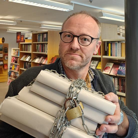 Hans Wendelboe Bøcher står med bogreoler i bagrunden. I hænderne foran sig, holder han en stak anonymiserede bøger, der er lænket sammen med jernkæder og hængelåse.