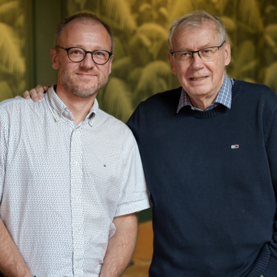 Dobbeltportræt af Hans Wendelboe Bøcher i hvid kortærmet skjorte. Til højre står filminstruktør Nils Malmros i mørkeblå striktrøje, med armen på Hans' skulder.