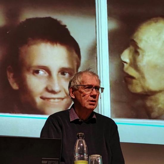 Nils Malmros står foran et lærred, hvor et billede fra filmen "Kundskabens træ", af en drengs ansigt kigger ud af et skab, ved siden af en gipsafstøbning.