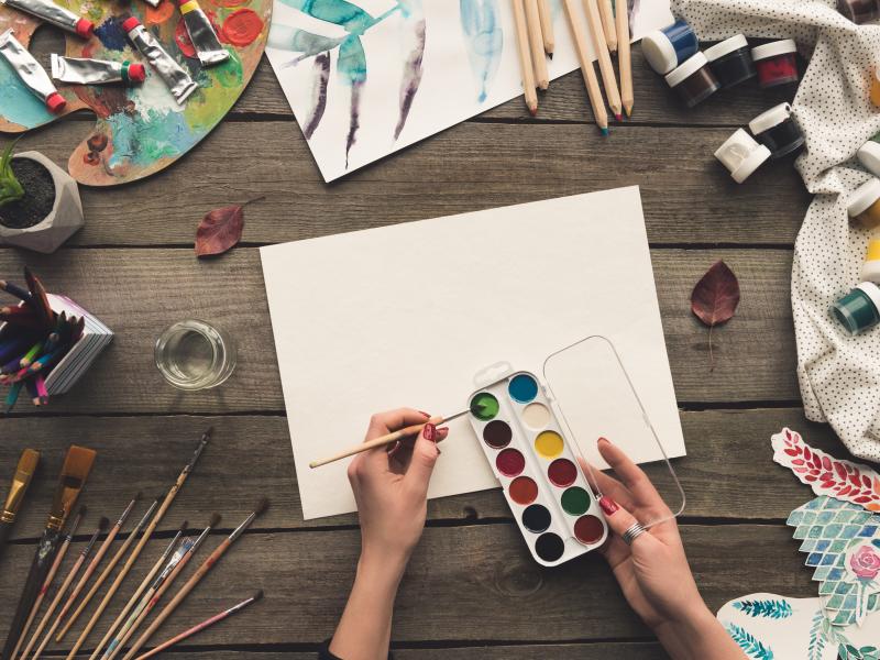 Viser et bord med materialer til tegning og maling og en hånde der maler med vandfarver