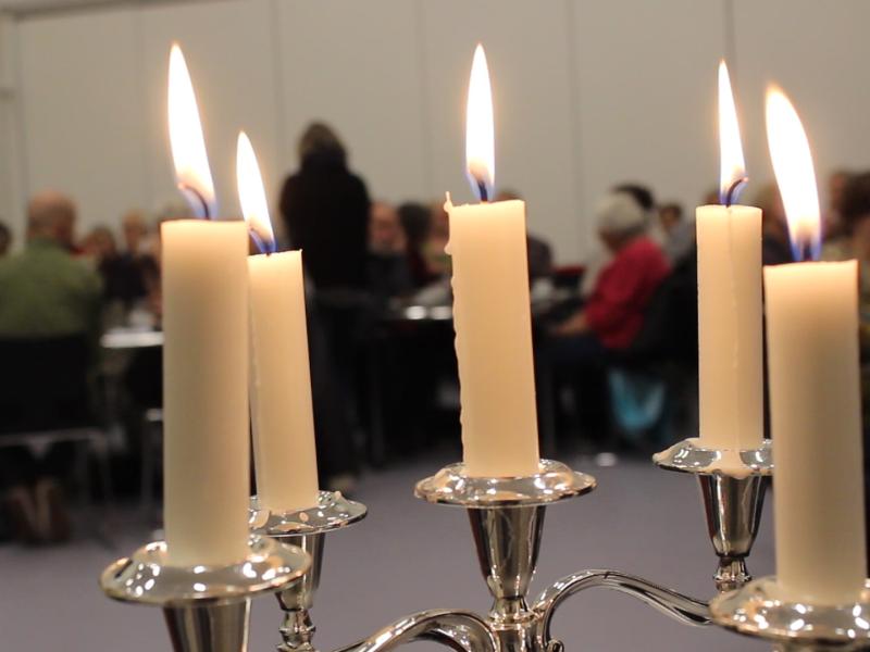 5 brændende stearinlys i forgrunden. Lyttende mennesker i baggrunden