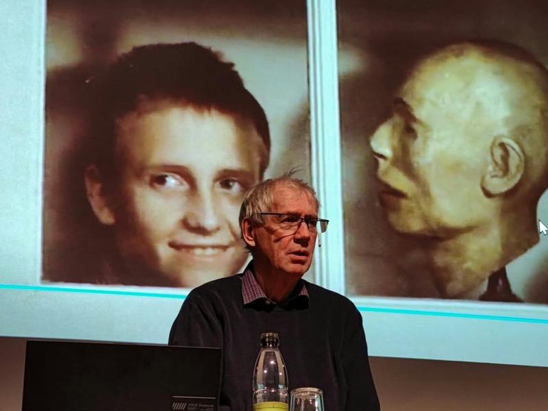Nils Malmros står foran et lærred, hvor et billede fra filmen "Kundskabens træ", af en drengs ansigt kigger ud af et skab, ved siden af en gipsafstøbning.