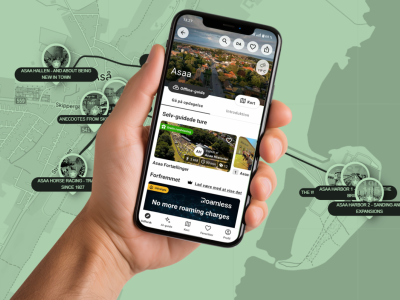 Hånd med smartphone, der viser app'en "SmartGuide" med historier om Asaa. Som baggrund ses et grøntonet kort over Asaa med hotspots.