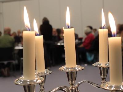 5 brændende stearinlys i forgrunden. Lyttende mennesker i baggrunden