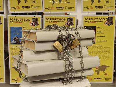 Stak af titelløse bøger, lænket sammen med jernkæder og hængelåse. I baggrunden hænger gule plakater med titler og tekster om bøger, der er, har været eller været forsøgt gjort forbudt. er