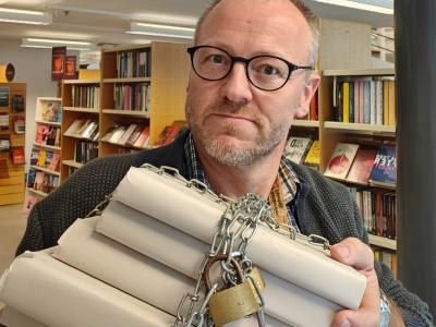 Hans Wendelboe Bøcher står med bogreoler i bagrunden. I hænderne foran sig, holder han en stak anonymiserede bøger, der er lænket sammen med jernkæder og hængelåse.