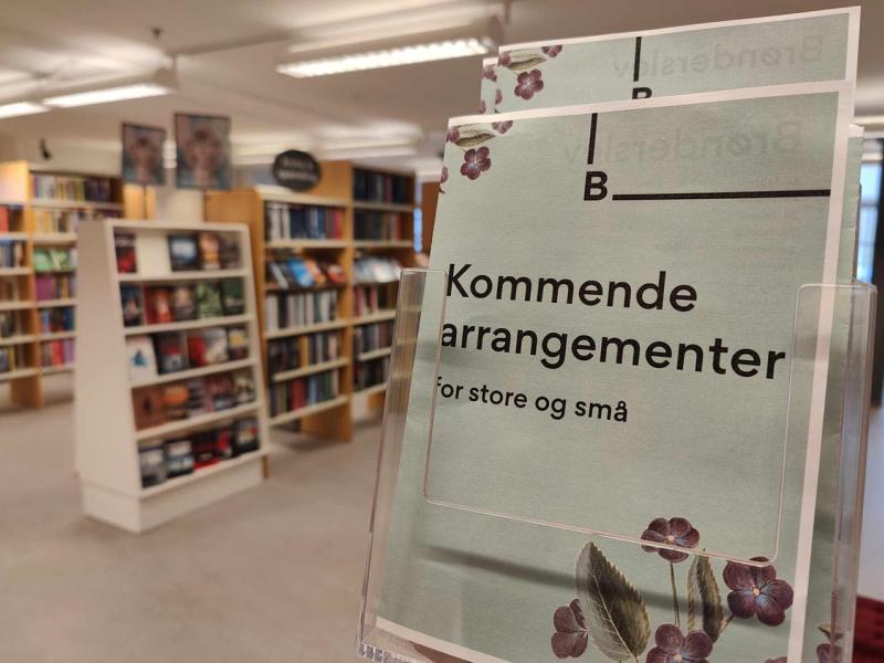 Stander med det trykte program for februar/marts 2025 på Brønderslev Bibliotek. I baggrunden ses sløret reolerne med krimier.