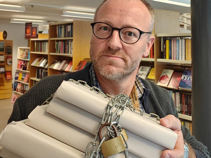 Hans Wendelboe Bøcher står med bogreoler i bagrunden. I hænderne foran sig, holder han en stak anonymiserede bøger, der er lænket sammen med jernkæder og hængelåse.