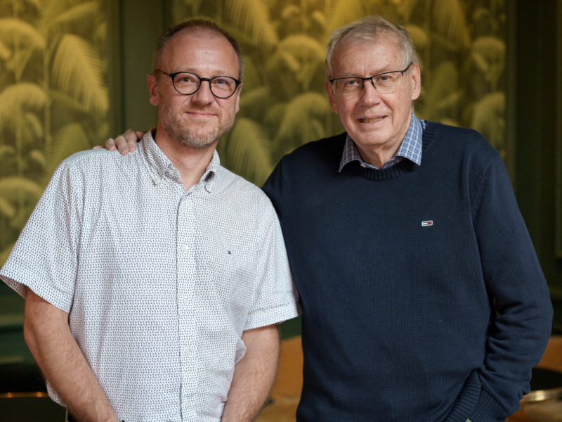 Dobbeltportræt af Hans Wendelboe Bøcher i hvid kortærmet skjorte. Til højre står filminstruktør Nils Malmros i mørkeblå striktrøje, med armen på Hans' skulder.
