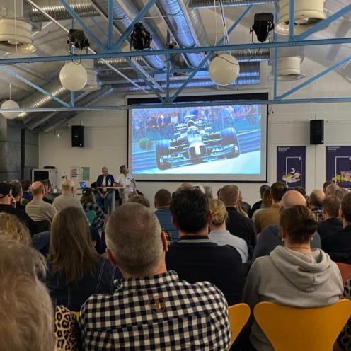 Publikum i salen på Brønderslev Bibliotek, set bagfra. På lærredet ses en formel1-racer.l1-arrangement 2025
