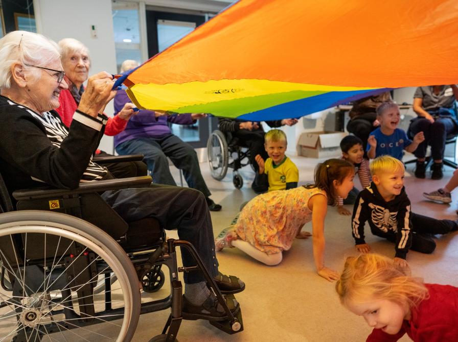 En glad, ældre biblioteksgæst i kørestol holder i en regnbuefarvet faldskærm. Under den kravler en masse børn.