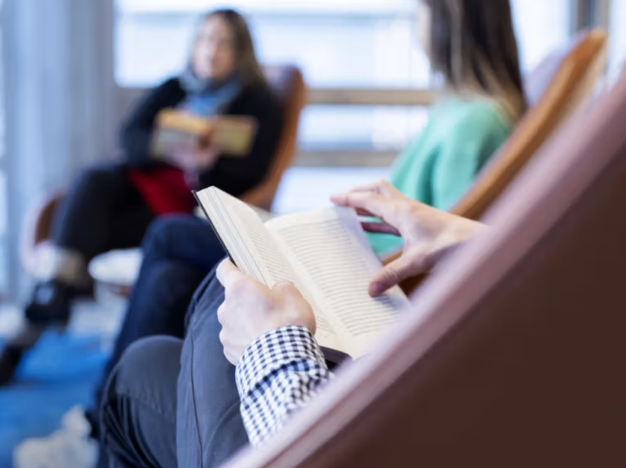 Nærbillede af hænder med bog. I baggrunden ses to personer, der lytter.