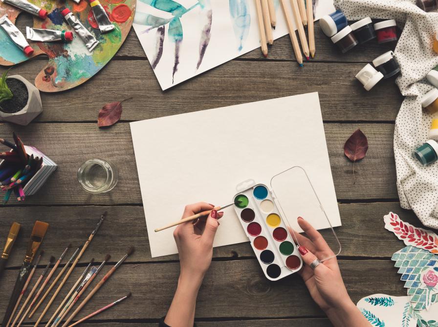 Viser et bord med materialer til tegning og maling og en hånde der maler med vandfarver