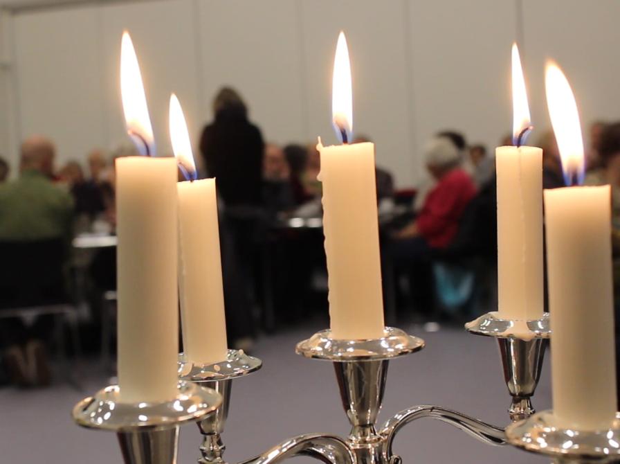 5 brændende stearinlys i forgrunden. Lyttende mennesker i baggrunden