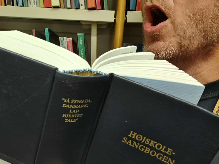 Nærbillede af Højskolesangbogen, med modificeret tekst på ryggen: "Så Syng Da, Brønderslev, Lad Hjertet Tale". I baggrunden mand, der synger og reol med bøger.