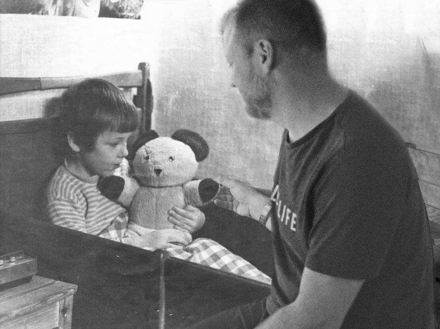 S/H-fotomanipulation af formidler Hans Wendelboe Bøcher som voksen, siddende på sengekanten, med ham selv som barn, der holder en bamse i favnen.