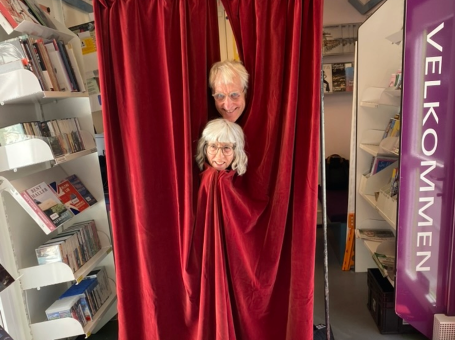 Billede fra Det mobile bibliotek hvor skuespillerne Birthe Rosenfeldt og Lars Olsen fra Aasen Teater står bag et rødt forhæng.