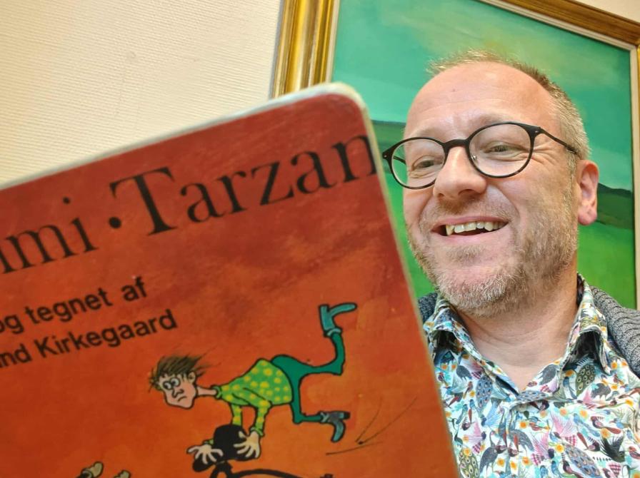 Formidler Hans Wendelboe Bøcher i spraglet skjorte læser Gummi-Tarzan. I baggrunden ses et grønt maleri.
